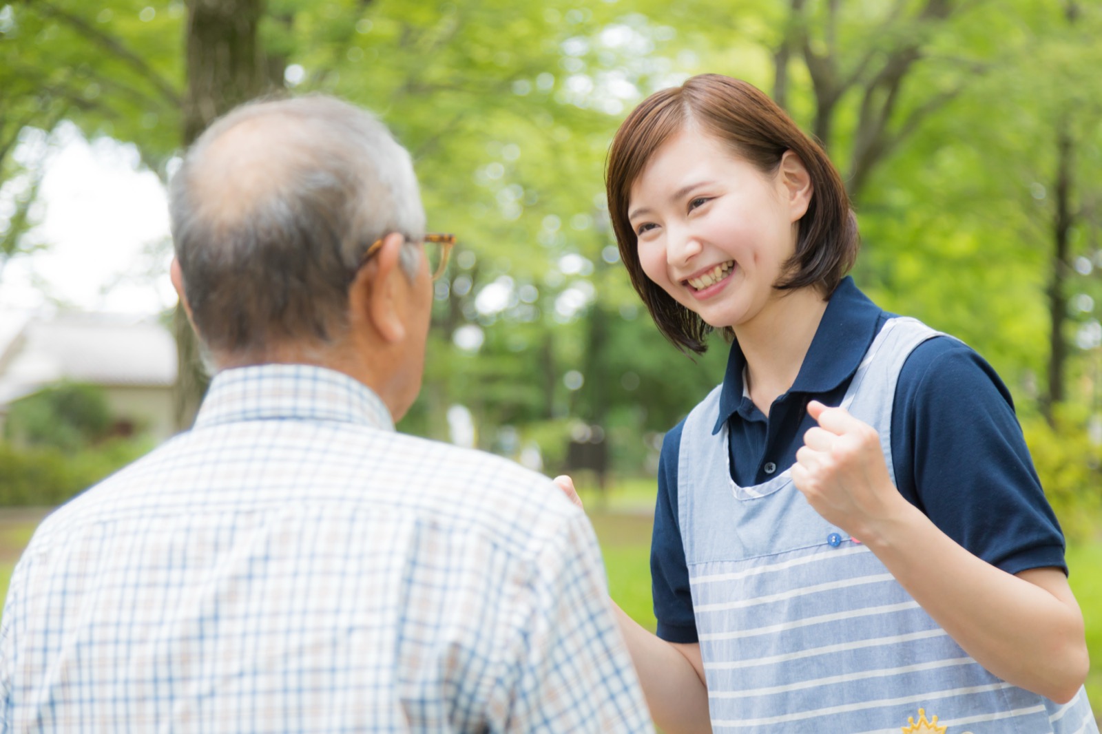 屋外で高齢の男性と向かい合い、笑顔で話しかけるエプロン姿の女性介護士
